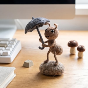 May include: A needle-felted brown ant figurine standing on a textured base, holding a small grey umbrella. The ant has black eyes and antennae. Two small needle-felted brown mushrooms are positioned nearby on the surface.