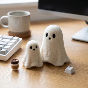 Puede incluir: Dos figuras de fantasmas de fieltro blanco con ojos de botón negros y bocas pequeñas. Una taza de café en miniatura y una tecla de teclado también están sobre la superficie de madera.