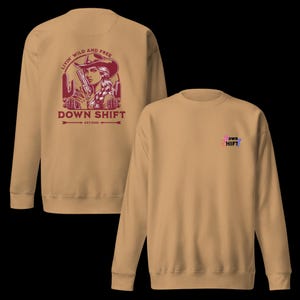Puede incluir: Sudadera color canela con un gráfico en la espalda que presenta a una vaquera con una pistola, cactus y el texto "LIVIN' WILD AND FREE" y "DOWN SHIFT". La parte delantera de la sudadera tiene un pequeño logotipo que dice "DOWN SHIFT".