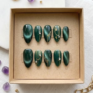 May include: A set of emerald green press-on nails in a cardboard box. Some nails have gold accents and rhinestones. The box is surrounded by amethyst stones, gold beads, and a gold ring.