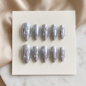 Pode incluir: Um conjunto de unhas postiças ovais iridescentes. Cada unha tem uma cor cinza-azulada clara e apresenta um design prateado de lua crescente ou estrela. As unhas estão dispostas em um cartão branco.