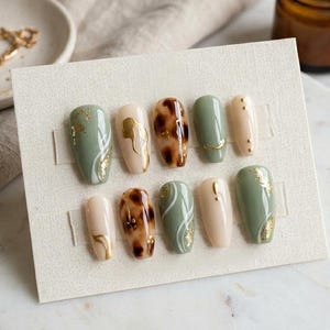 May include: A set of ten press-on nails in various designs. The nails feature colors like sage green, nude, and tortoiseshell, with gold foil and line accents. The nails are displayed on a white card, with a candle and jewelry in the background.