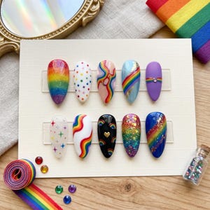 Pode incluir: Um conjunto de dez unhas postiças com vários designs, incluindo gradientes de arco-íris, bolinhas e padrões abstratos. As unhas são exibidas em um cartão branco, com uma fita de arco-íris e glitter por perto. A paleta de cores é vibrante, com várias cores.