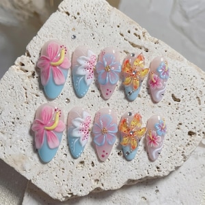 Könnte beinhalten: Ein Set aus zehn künstlichen Nägeln mit floralem Design. Jeder Nagel zeigt eine andere Blume, darunter rosa Hibiskus, weiße Lilien und blaue und gelbe Blüten. Die Nägel haben eine hellblaue Basis und sind mit Strasssteinen verziert.