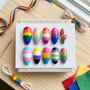 以下が含まれることがあります： LGBTQ+プライドフラッグをそれぞれ異なるデザインで表現した、10本のつけ爪。デザインには、虹色のストライプ、グリッター、旗のパターンが含まれています。つけ爪は白いカードに展示されており、虹色のリボンとビーズのアクセントが付いています。