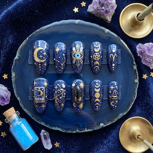 Op de afbeelding: Een set van tien marineblauwe nepnagels met gouden hemelse ontwerpen. Elke nagel heeft een ander ontwerp, waaronder manen, sterren, sterrenbeelden en een oog. De nagels worden gepresenteerd op een blauwe agaat schijf.