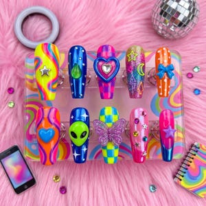 May include: A collection of ten long, coffin-shaped press-on nails. Each nail features a unique design, including swirls, hearts, stars, and an alien. Colors include neon yellow, blue, pink, orange, and green. A disco ball and a smartphone are in the background.