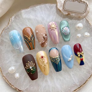 May include: A collection of ten decorated artificial nails in various colors and designs. The nails feature embellishments like pearls, a sun, a seashell, and floral motifs. The color palette includes blue, brown, pink, and gold, with a pearlescent finish.
