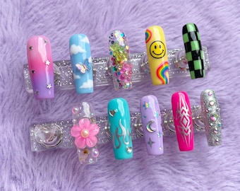 May include: A collection of ten long, rectangular press-on nails with various designs. The designs include a smiley face, clouds, butterflies, stars, flames, and glitter. Colors include pink, purple, blue, green, and yellow. The nails are displayed on a clear, sparkly holder.