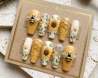 May include: A set of press-on nails with a bee and floral theme. The nails feature yellow, white, and clear designs, including bees, sunflowers, honeycomb, and daisies. The nails are displayed on a brown card.