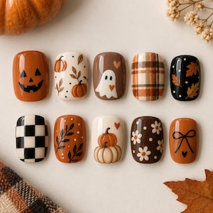 May include: A set of ten press-on nails featuring autumn-themed designs including a jack-o'-lantern, pumpkins, a ghost, plaid patterns, black and white checkers, fall leaves, white daisies, and a black bow on orange, brown, black, and cream backgrounds.
