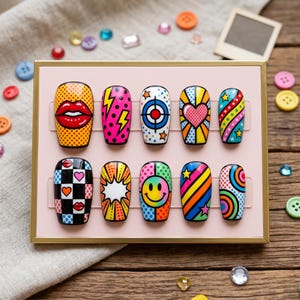 Könnte beinhalten: Ein Set aus zehn farbenfrohen Press-on-Nägeln mit verschiedenen Pop-Art-Designs. Die Nägel zeigen auffällige Grafiken wie Lippen, Blitze, Herzen und Smiley-Gesichter. Die Nägel sind in einem rosa Display-Case angeordnet.