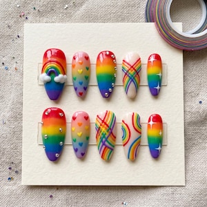 Pode incluir: Um conjunto de dez unhas postiças com tema arco-íris. Cada unha apresenta um design diferente, incluindo arco-íris, corações e estrelas. As unhas estão dispostas em um cartão branco, com um rolo de fita colorida no fundo.