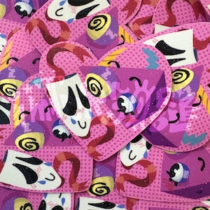 May include: A collection of heart-shaped stickers featuring a repeating pattern of a white mask with black eyes and a blue tear, a yellow swirl, and a red ribbon on a pink background with a dotted texture and a subtle glittery finish.