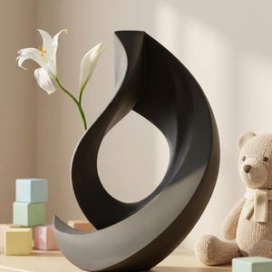 Può includere: Un vaso in ceramica nero opaco con una silhouette curva, astratta e a forma di anello aperto. Un singolo giglio bianco con steli verdi è posizionato all'interno del vaso. Vicino ci sono diversi piccoli blocchi di legno pastello e un orsacchiotto lavorato a maglia.