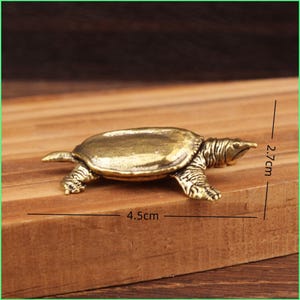 Może przedstawiać: Mała, szczegółowa figurka żółwia z mosiądzu, o długości 4,5 cm i wysokości 2,7 cm, prezentowana na drewnianej powierzchni. Żółw ma teksturowany pancerz i nogi, z długim, spiczastym pyskiem. Mosiądz ma ciepłe, antyczne wykończenie.
