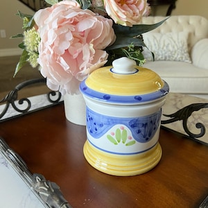 May include: A ceramic canister with a lid, featuring a yellow base and top, a blue band with dots, and a central white section decorated with blue brushstrokes and small green and pink floral-like motifs painted on the exterior surface.
