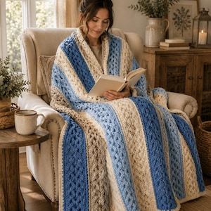 Puede incluir: Una manta de ganchillo con rayas verticales en tonos crema, azul claro y azul oscuro. La manta presenta un patrón texturizado de punto trenzado en relieve, con secciones de colores sólidos alternadas y detalles de trenzas.