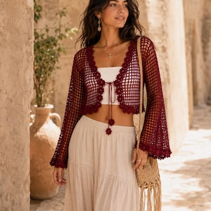 May include: A deep burgundy open-front crochet cardigan featuring a grid pattern, long flared sleeves, and a scalloped floral trim along the edges. The garment includes thin matching ties at the center with small crocheted flower accents at the ends.