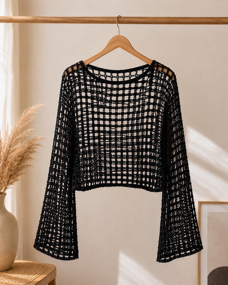 May include: A black long-sleeve top featuring an open-knit crochet construction with a grid-like pattern. The garment has a wide, rounded neckline and flared sleeves that extend from the shoulders, creating a loose, airy silhouette throughout.