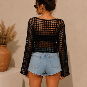 May include: A black long-sleeve crochet top featuring an open-knit grid pattern, a wide neckline, and flared sleeves, paired with light blue denim shorts that have a frayed hemline and a classic five-pocket design with metal rivets.