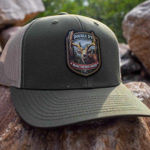 May include: An olive green baseball cap with a tan mesh back and a structured front panel. A shield-shaped patch on the front features a deer illustration with the text "DOUBLE D'S" and "MAKE THE BEST RACKS" printed in white lettering.