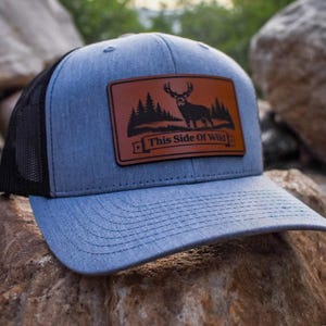 May include: A heather gray baseball cap with a black mesh back and a rectangular brown leather patch on the front. The patch features a black graphic of a deer in a forest with the text This Side Of Wild printed in a banner at the bottom.