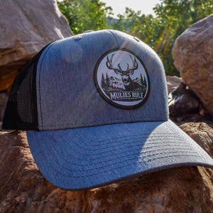 May include: A heather gray baseball cap with a black mesh back and a circular patch on the front. The patch features a black and white illustration of a deer with large antlers in front of mountains and trees, with the text MULIES RULE.