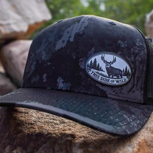 May include: A black and dark gray camouflage patterned trucker hat with a black mesh back. An oval patch on the front features a white background with a black silhouette of a deer standing in a forest and the text This Side of Wild.