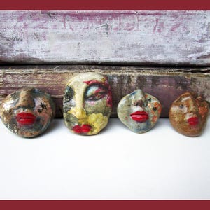 May include: Four small, hand-sculpted ceramic face ornaments with glossy red lips. Each piece features unique, textured surfaces in shades of brown, gray, and cream, with subtle painted details including small black hearts and floral-like patterns.