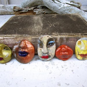 May include: Five small, hand-sculpted clay faces arranged in a row. Each face features distinct colors and expressions, including metallic gold, copper with blue lips, a weathered beige with red lips, a glossy red, and a yellow with painted details.