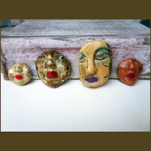 May include: Four small, hand-sculpted ceramic face ornaments arranged in a row. Each piece features distinct facial features, including painted eyes and lips in red or purple. The faces vary in color, ranging from cream and brown to yellow and orange.