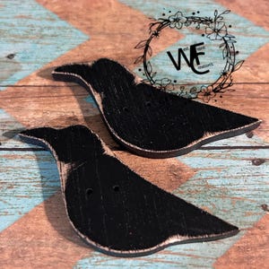 May include: Two flat, bird-shaped wooden cutouts painted matte black with distressed, natural wood-toned edges. Each bird features two small circular holes in the center. A circular logo with the text "WF est. front reation's" is visible.