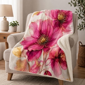 Peut inclure: Une couverture douillette ornée d'un grand motif floral rose et doré. La couverture a un envers en sherpa et est drapée sur un fauteuil de couleur neutre. Le motif floral évoque une peinture à l'aquarelle.