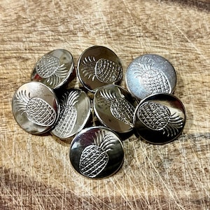 May include: A group of silver metal buttons with a pineapple design. The buttons are arranged in a pile on a wooden surface.