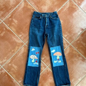 May include: Blue denim jeans with two patches on the knees. Each patch features a mushroom, clouds, and a red bow.