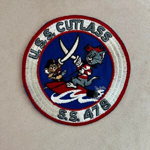 May include: A blue and red embroidered patch with a white border. The patch features a cartoon cat and mouse fighting with swords. The text "U.S.S. CUTLASS S.S. 478" is around the edge of the patch.