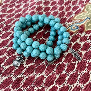 May include: A turquoise beaded bracelet with silver charms, including an owl and a feather. The bracelet is made of round, textured beads and is displayed on a patterned red and cream fabric.