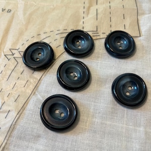 May include: Six dark blue buttons with a marbled effect, each featuring two holes for sewing. The buttons are arranged on a light-colored fabric, possibly linen, with a pattern piece visible in the background.