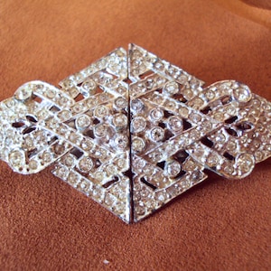May include: A silver-toned brooch with a geometric design, featuring a large number of clear rhinestones.