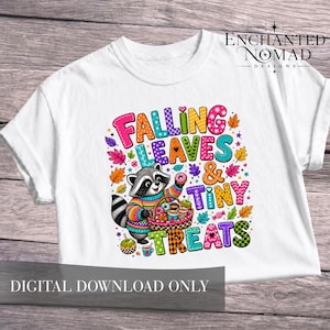 May include: A white t-shirt featuring a colourful graphic of a raccoon wearing a striped jumper and holding a basket of doughnuts and apples. The text reads FALLING LEAVES & TINY TREATS, surrounded by vibrant autumn leaves and small stars.