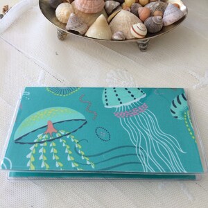 May include: A teal blue notebook with a clear plastic cover. The cover features a colorful illustration of jellyfish with pink, yellow, and green accents. The notebook is closed and sitting on a white surface.