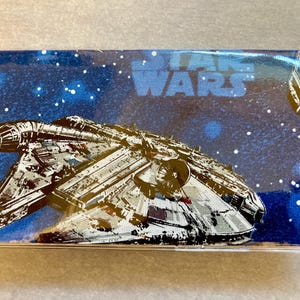 Puede incluir: Un artículo rectangular con temática de Star Wars, que muestra la nave espacial Halcón Milenario sobre un fondo azul estrellado. Las palabras "STAR WARS" son visibles en azul. El artículo está en plástico transparente.