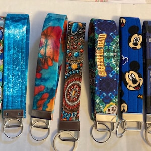 May include: Eight colorful fabric keychains with metal key rings. The keychains feature various designs, including a blue and white stone pattern, a blue and white wave pattern, a blue and red tie-dye pattern, a red and gold beaded pattern, a blue and green tie-dye pattern with the text "Grateful Dead", a blue keychain with a Mickey Mouse character, and a blue and black keychain with a repeating pattern of a blue police box.