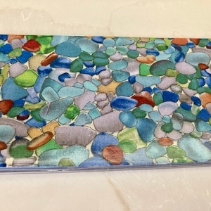 May include: A close-up of a fabric with a pattern of colorful, smooth, and rounded stones. The stones are various shades of blue, green, brown, and purple. The background is a light cream color.