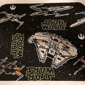 Könnte beinhalten: Schwarzes Mauspad mit Star Wars-Motiv. Das Design zeigt den Millennium Falken, X-Wing-Jäger und das Star Wars-Logo in Gelb. Der Hintergrund ist ein Sternenhimmel mit Symbolen der Rebellenallianz.