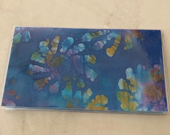 May include: A checkbook cover with a vibrant, abstract batik design. The cover features a clear plastic sleeve and a blue background with colorful, organic shapes in shades of yellow, purple, and teal. The overall design is artistic and eye-catching.