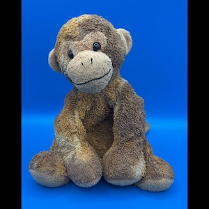May include: A plush monkey toy with textured brown and tan fur. The stuffed animal features a light tan face, dark brown ears, and black plastic eyes. It has a stitched black mouth line and is seated in an upright position with long arms.