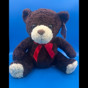 May include: A plush teddy bear with dark brown fur, a cream-coloured muzzle, and cream-coloured paw pads. The bear features dark, round eyes, a brown nose, and a bright red ribbon tied in a bow around its neck. A small product tag is attached to the ear.