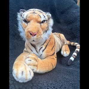 May include: A plush tiger toy featuring orange fur with black stripes, a white underbelly, and white fur around the cheeks. The tiger has a pink nose, amber-coloured eyes, and is depicted in a resting position with its front paws crossed together.
